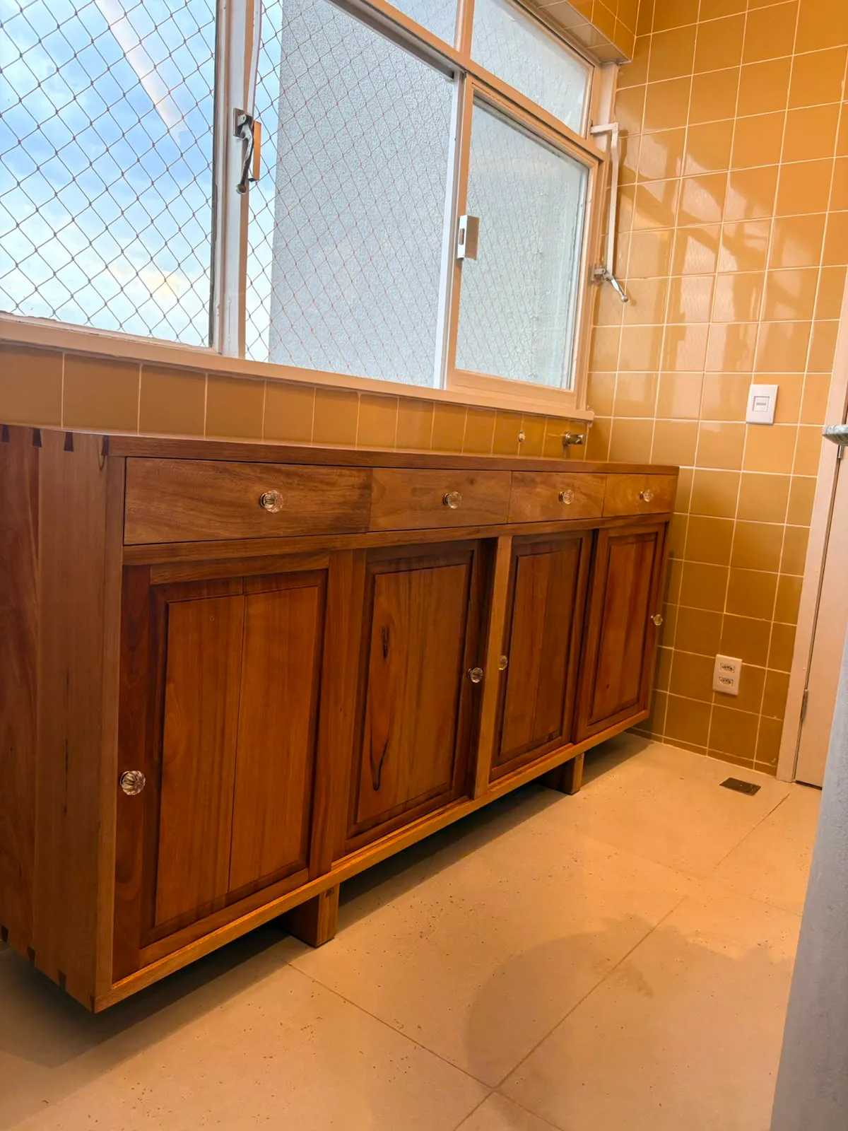 Buffet de madeira maciça encostado em parede de azulejo amarelo, com quatro gavetas de puxadores de vidro, quatro portas almofadadas e encaixes de rabo-de-andorinha aparentes no canto do tampo.