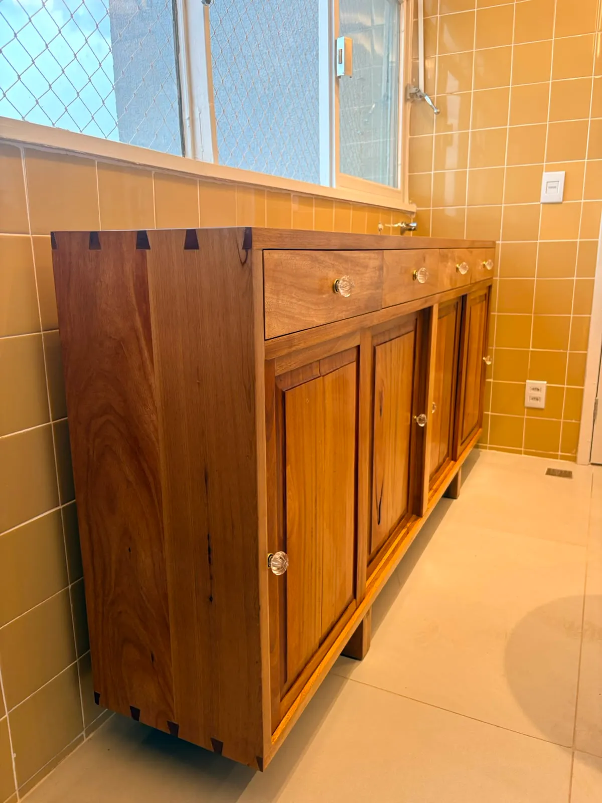Buffet de madeira maciça encostado em parede de azulejo amarelo, com quatro gavetas de puxadores de vidro, quatro portas almofadadas e encaixes de rabo-de-andorinha aparentes no canto do tampo. — foto 2