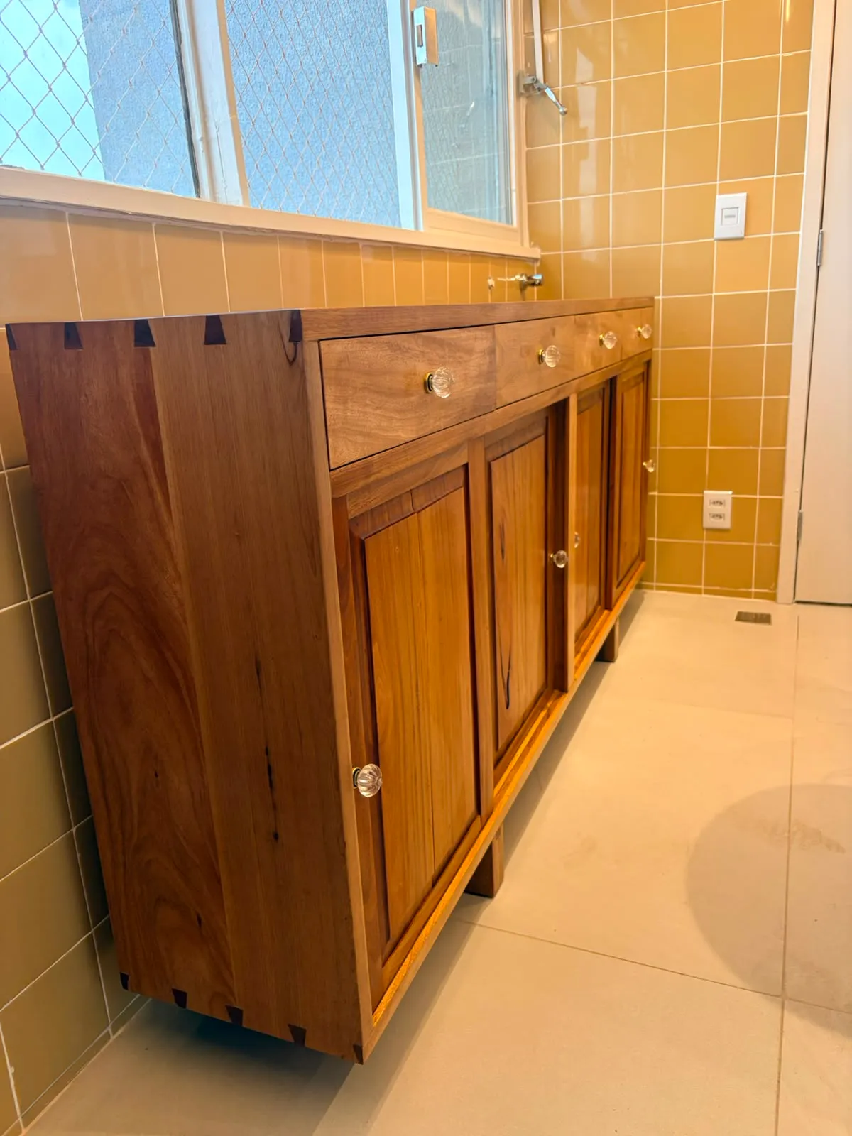 Buffet de madeira maciça encostado em parede de azulejo amarelo, com quatro gavetas de puxadores de vidro, quatro portas almofadadas e encaixes de rabo-de-andorinha aparentes no canto do tampo. — foto 3