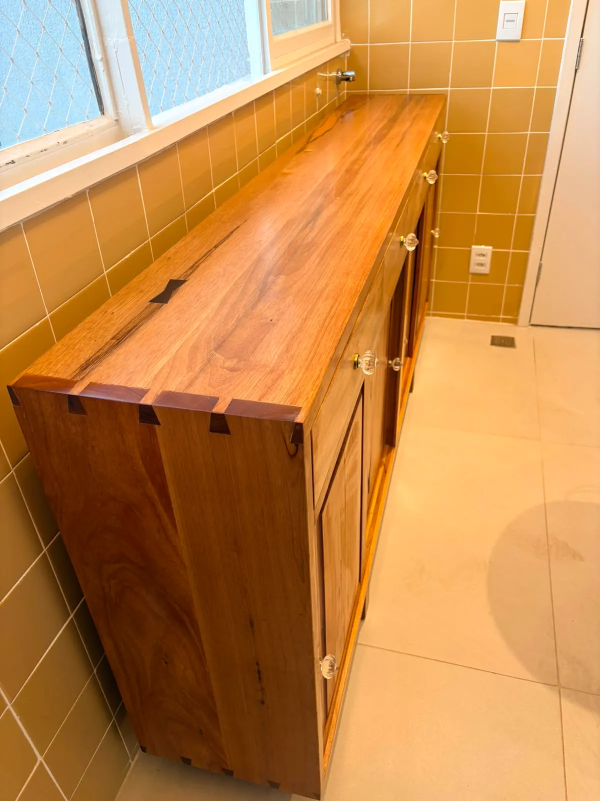 Buffet de madeira maciça encostado em parede de azulejo amarelo, com quatro gavetas de puxadores de vidro, quatro portas almofadadas e encaixes de rabo-de-andorinha aparentes no canto do tampo. — foto 4