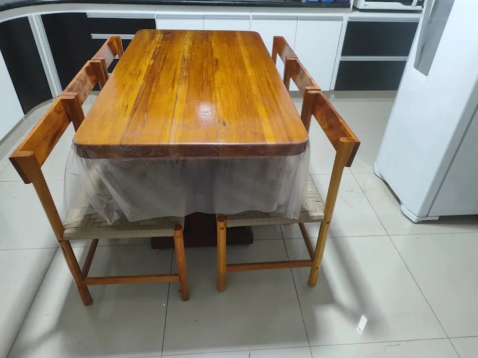 Mesa retangular de madeira maciça com cantos arredondados e duas cadeiras de assento trançado, numa cozinha de armários brancos.