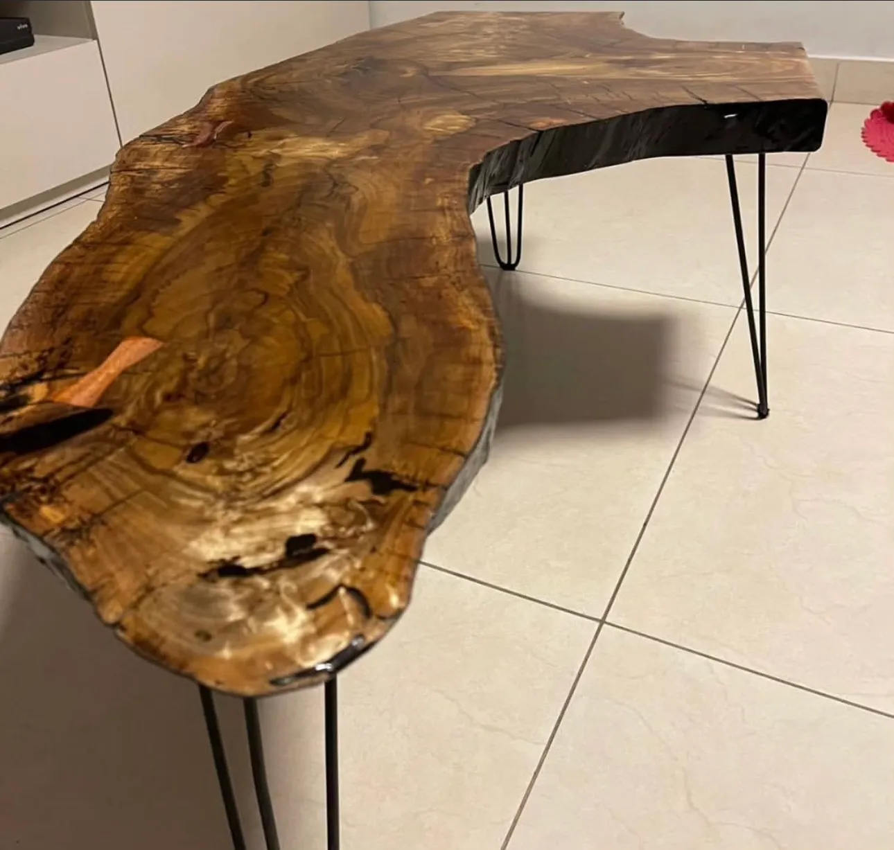 Mesa de centro feita de uma prancha de madeira de borda viva e formato irregular, apoiada em pés finos de ferro preto, numa sala de piso claro.