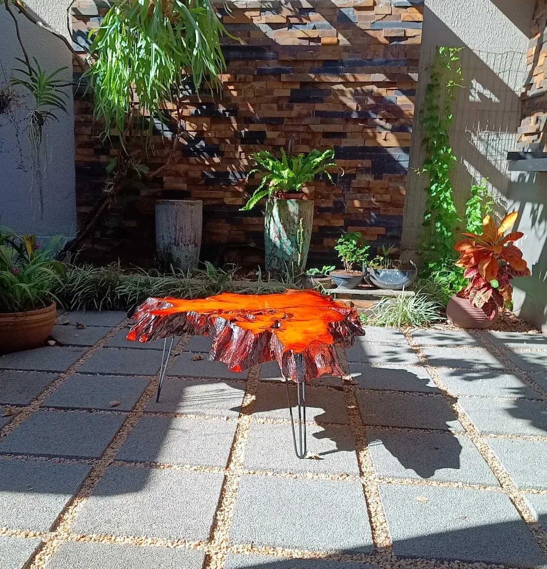 Mesa de centro feita de uma fatia de tronco com resina laranja preenchendo as fendas, apoiada em pés finos de ferro, num jardim com parede de pedra.