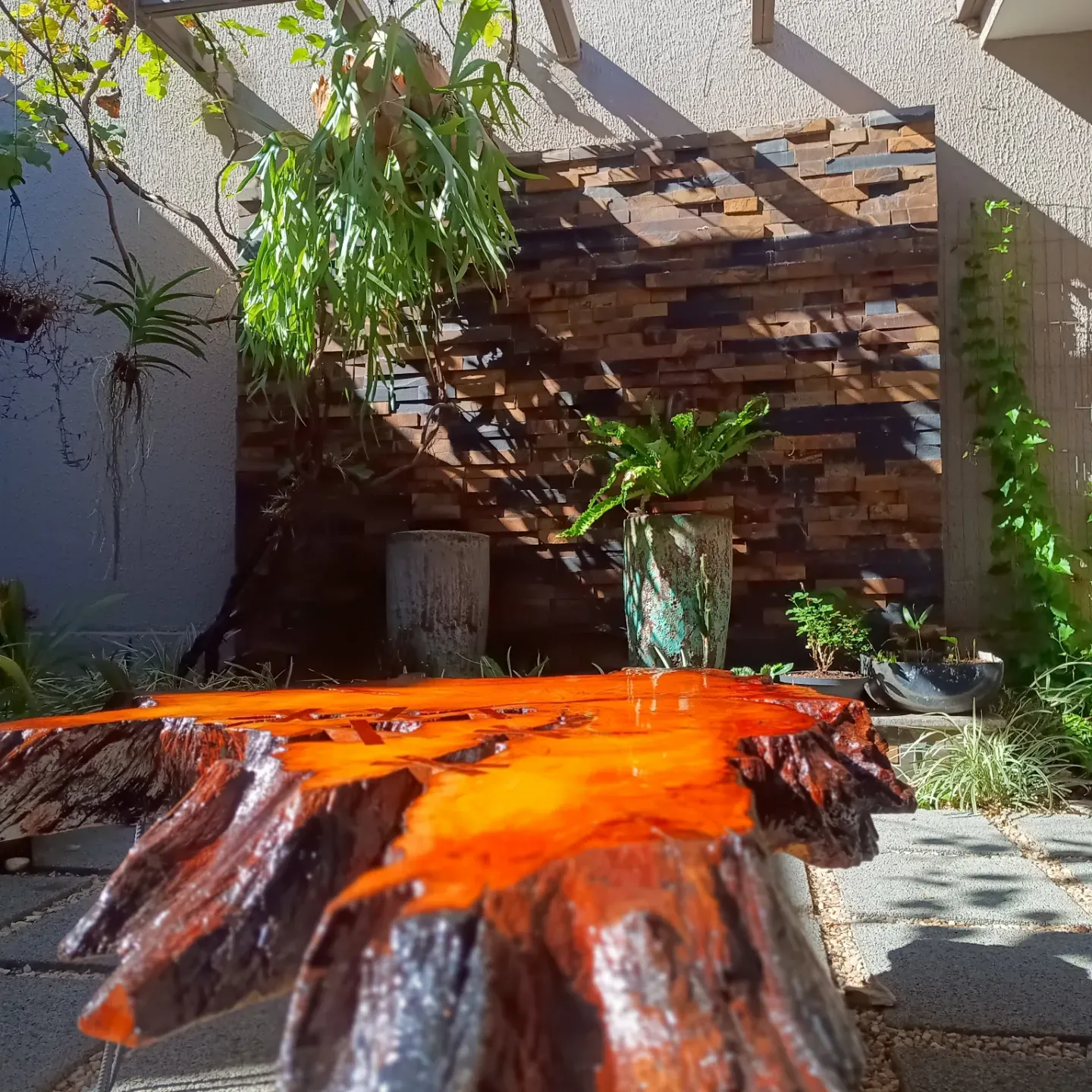 Mesa de centro feita de uma fatia de tronco com resina laranja preenchendo as fendas, apoiada em pés finos de ferro, num jardim com parede de pedra. — foto 2