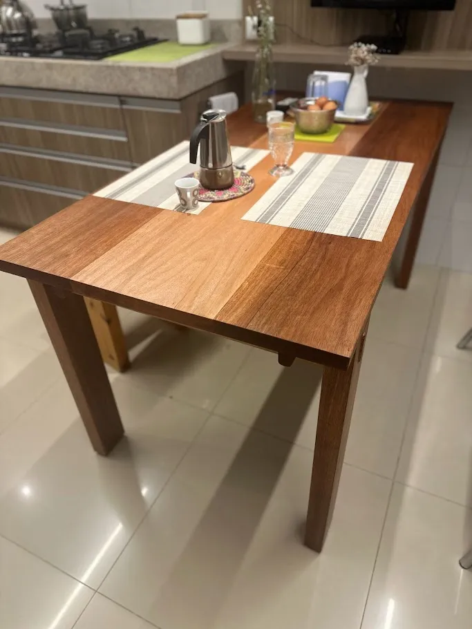 Mesa de cozinha em madeira maciça com quatro pernas e cavilhas aparentes nas travessas, posta com jogo americano e cafeteira italiana.