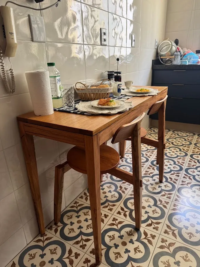 Mesa estreita de madeira maciça encostada na parede de uma cozinha, posta para duas pessoas, com duas cadeiras de assento redondo e encosto curvo.