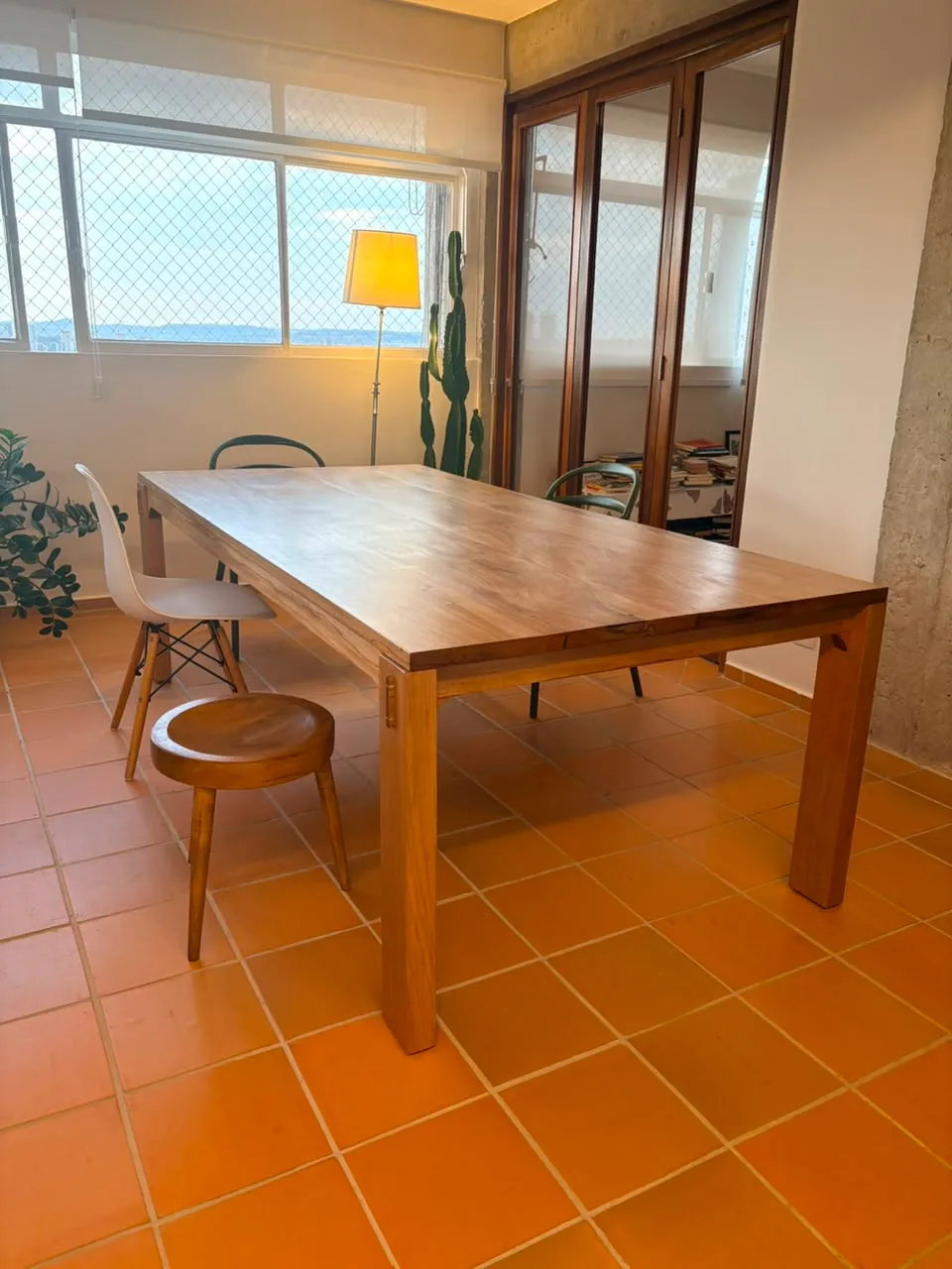 Mesa de jantar retangular em jequitibá-rosa, de pernas quadradas com cavilhas escuras aparentes, sobre piso de cerâmica laranja, entre uma janela e uma parede de concreto aparente.