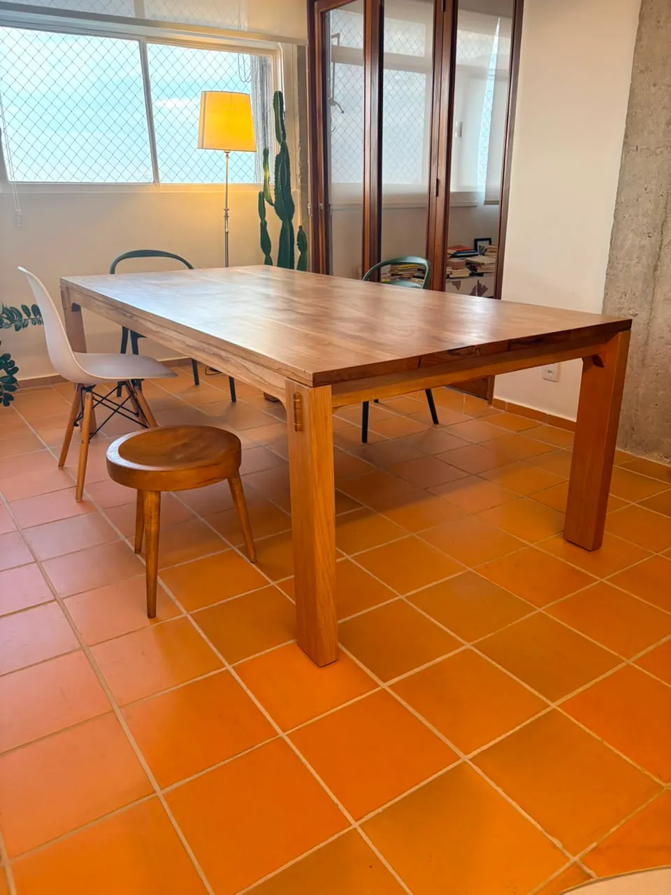 Mesa de jantar retangular em jequitibá-rosa, de pernas quadradas com cavilhas escuras aparentes, sobre piso de cerâmica laranja, entre uma janela e uma parede de concreto aparente. — foto 2