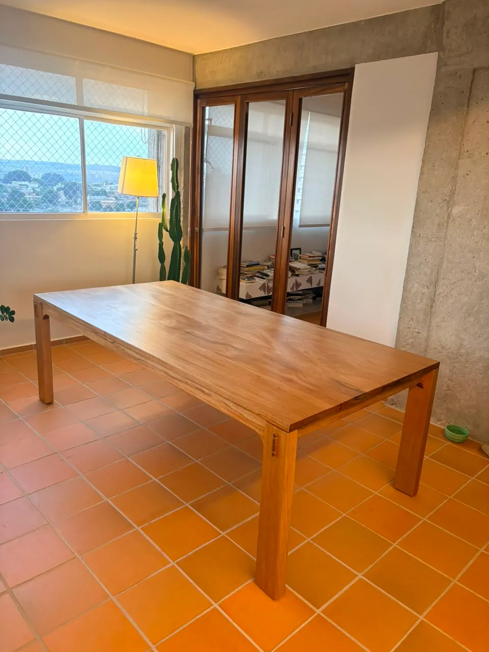 Mesa de jantar retangular em jequitibá-rosa, de pernas quadradas com cavilhas escuras aparentes, sobre piso de cerâmica laranja, entre uma janela e uma parede de concreto aparente. — foto 3