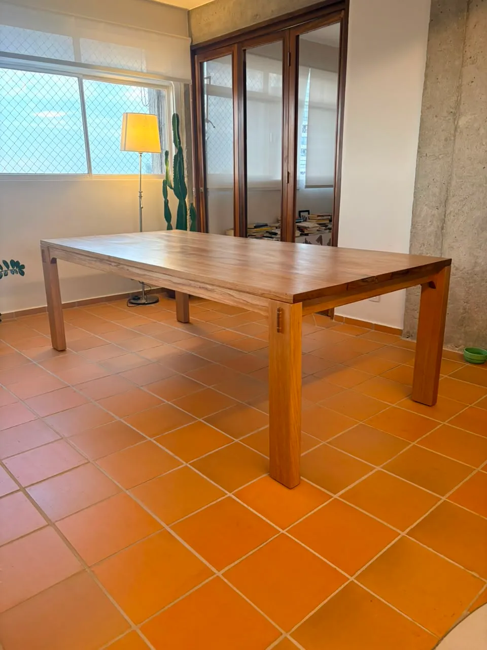 Mesa de jantar retangular em jequitibá-rosa, de pernas quadradas com cavilhas escuras aparentes, sobre piso de cerâmica laranja, entre uma janela e uma parede de concreto aparente. — foto 4