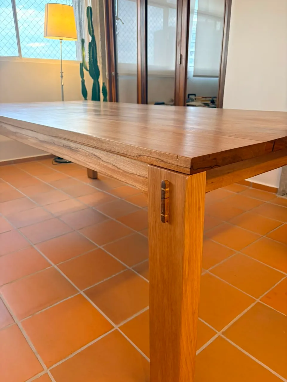 Mesa de jantar retangular em jequitibá-rosa, de pernas quadradas com cavilhas escuras aparentes, sobre piso de cerâmica laranja, entre uma janela e uma parede de concreto aparente. — foto 5
